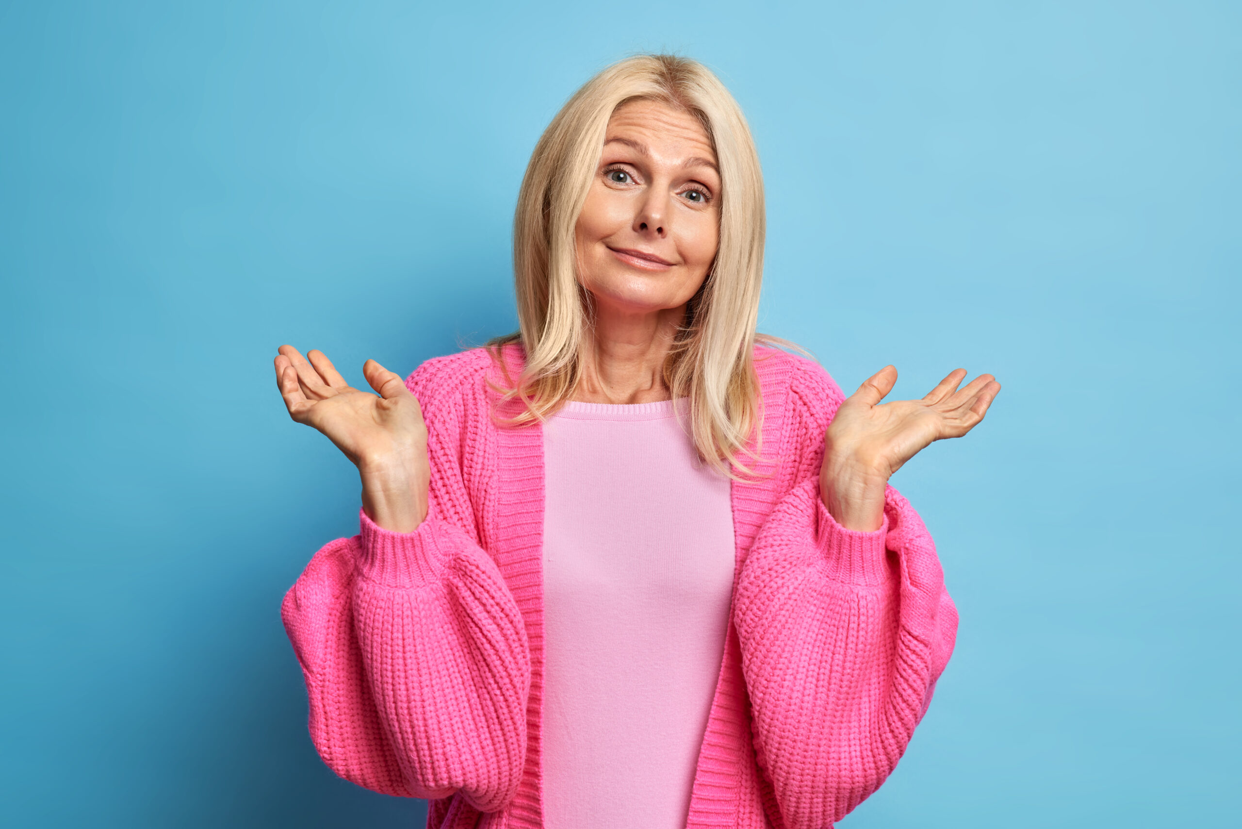 Puzzled perplexed woman shrugs shoulders looks doubtful and hesitant considers while makes decision dressed in pink clothes isolated over blue background. Unaware clueless senior lady gestures unsure