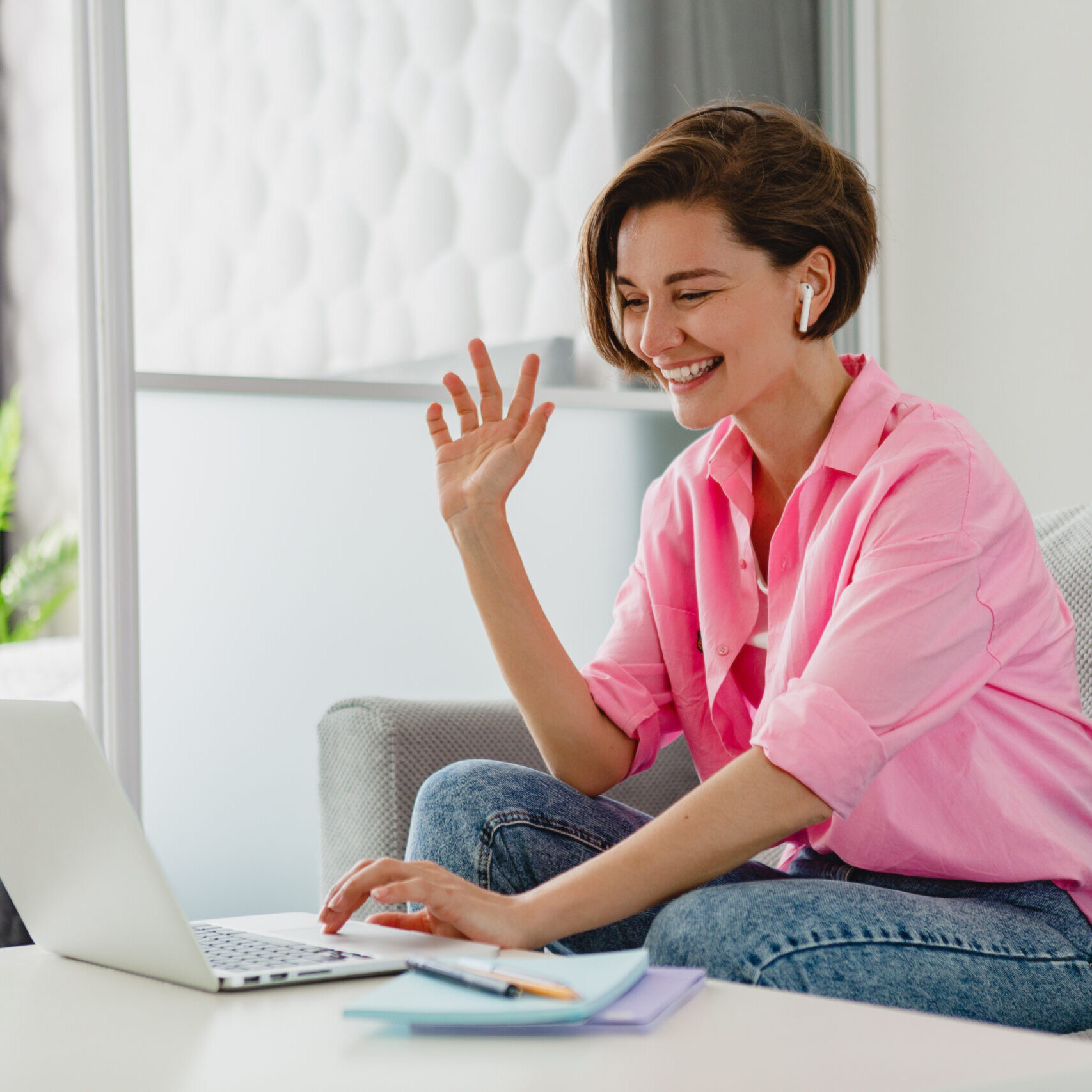 attractive smiling woman in pink shirt sitting relaxed on sofa at home at table working online on laptop from home freelancer, social distancing self isolation communication digital conference